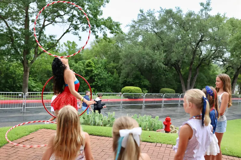 A hula hooper balancing a hoop on her face while kids watch